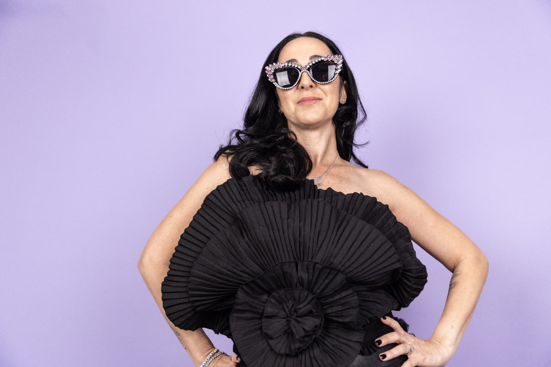 Rachel Sobel wearing jeweled sunglasses and a black ruffled dress against a purple backdrop
